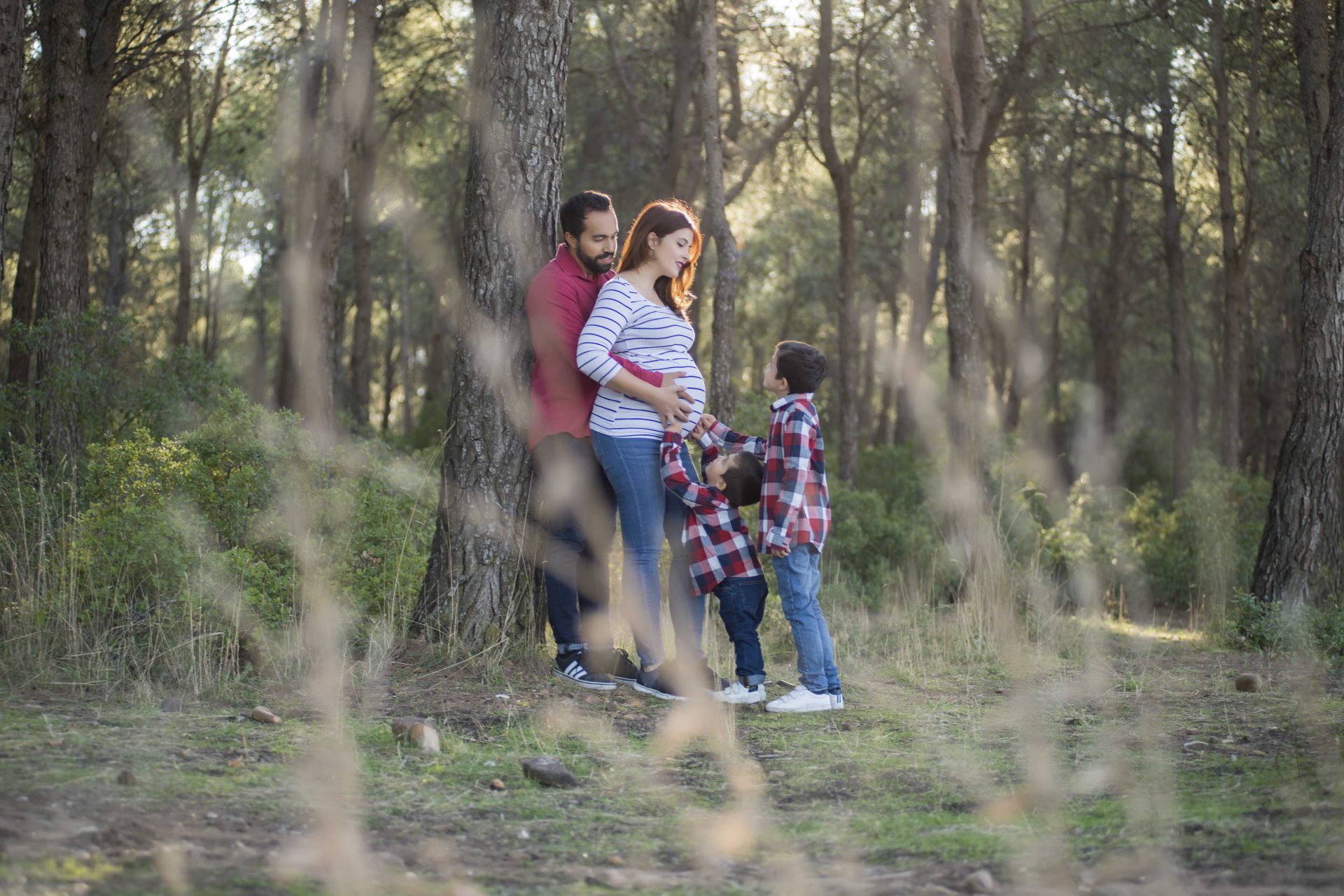 Fotografía de embarazada con su pareja en el bosque apoyados en un pino y sus dos otros hijos