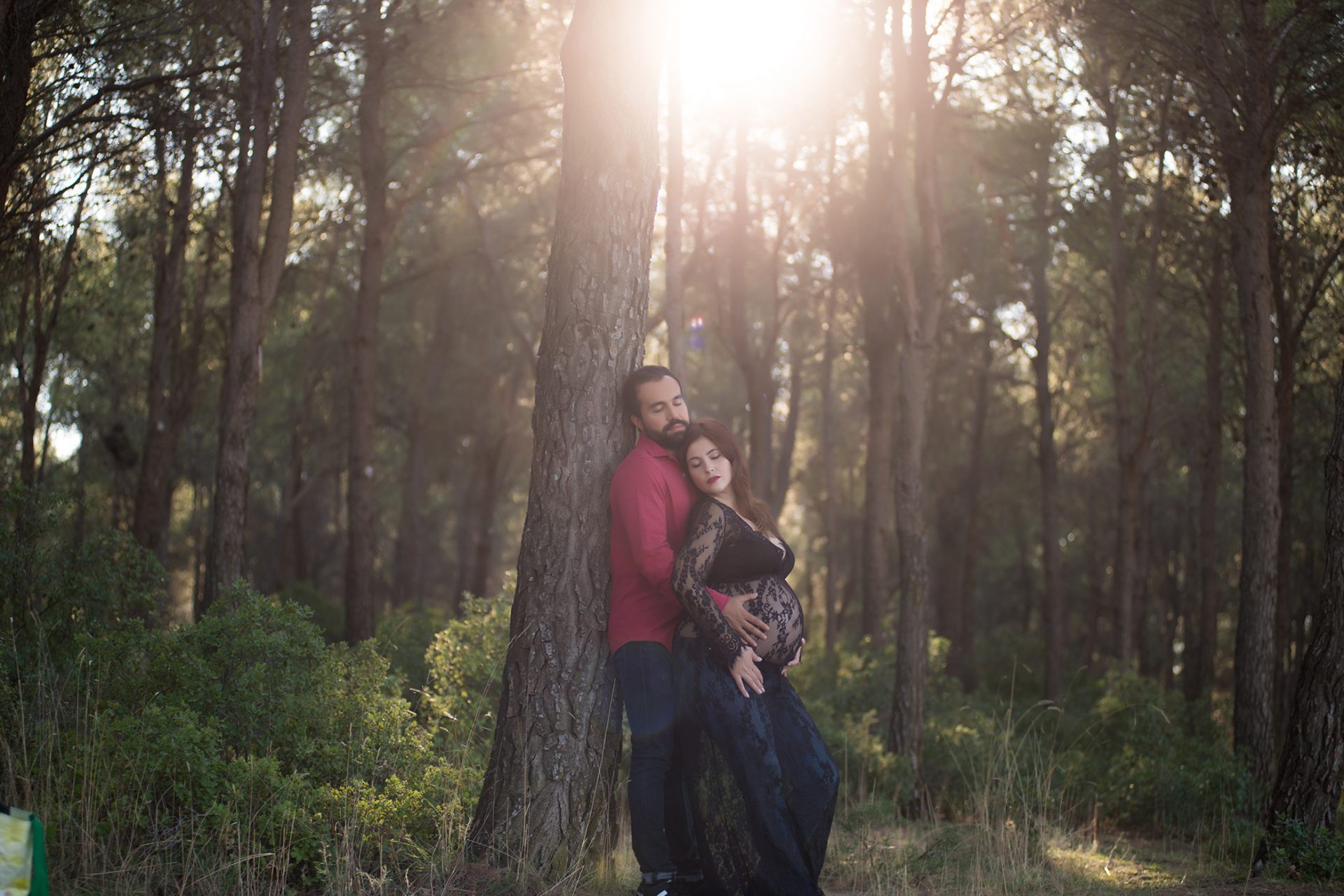 Fotografía de embarazada con su pareja en el bosque apoyados en un pino