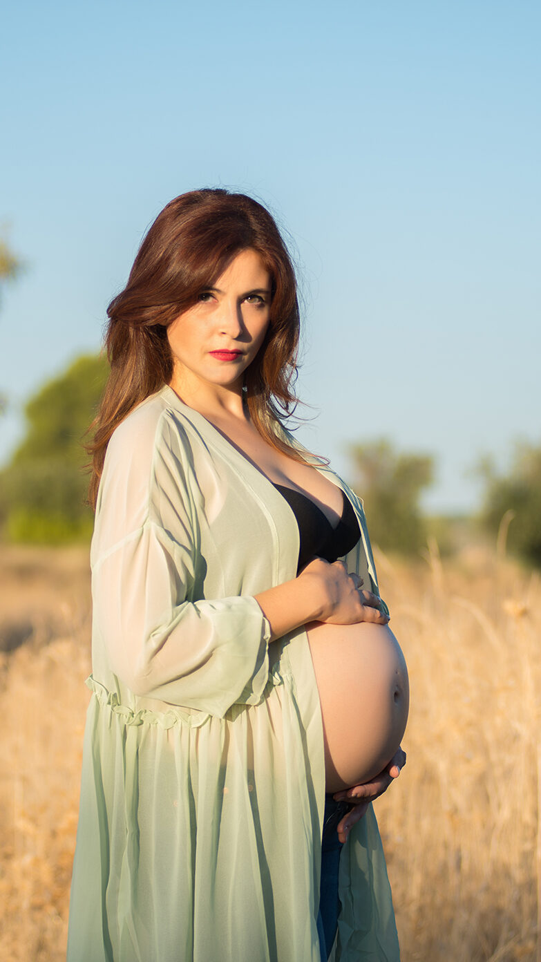 Fotografía de mujer embarazada en el campo con camisón verde mirando a cámara